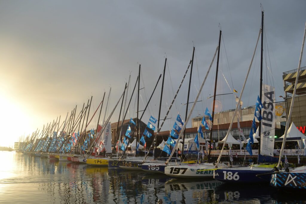 plusieurs bateaux de voile sont au ponton du village de la transat jacques vabres, le soleil se levant en arrière plan.