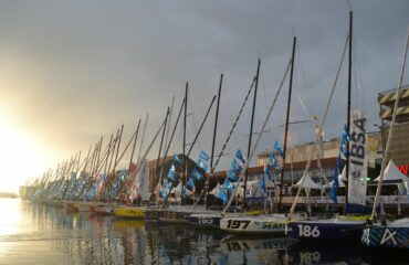 plusieurs bateaux de voile sont au ponton du village de la transat jacques vabres, le soleil se levant en arrière plan.