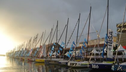 plusieurs bateaux de voile sont au ponton du village de la transat jacques vabres, le soleil se levant en arrière plan.